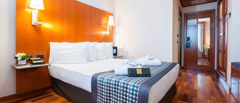 Habitación de hotel con cabecera de madera, cama con ropa blanca, mesita de noche con lámpara, una computadora portátil y una planta.