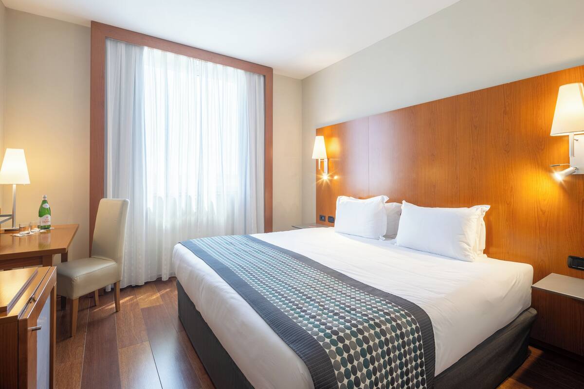 A hotel room with a large bed, a desk with a lamp, a chair, and a window with curtains.