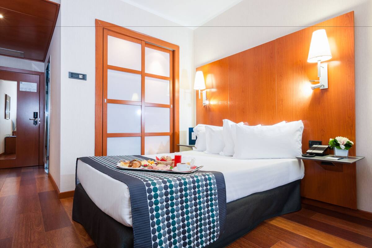 A hotel room with a bed, a tray of breakfast, a nightstand with a lamp, and a wooden door.