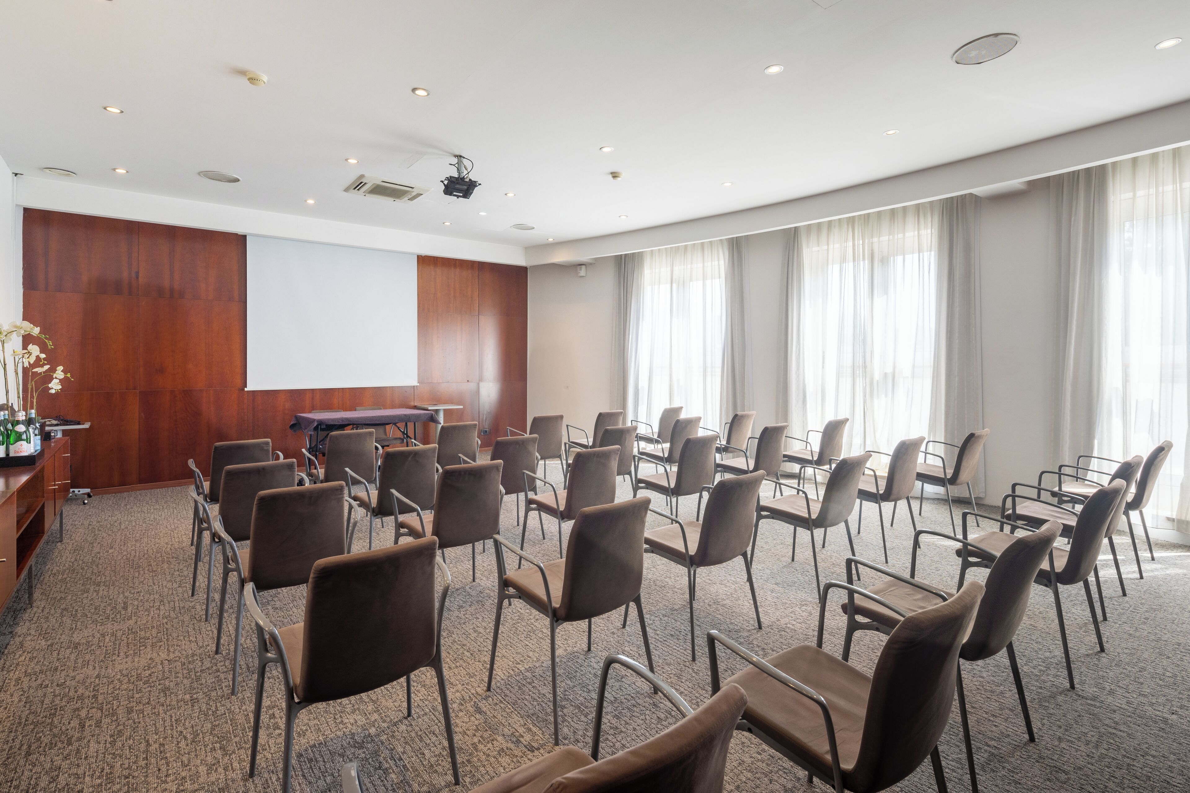 Un salón de conferencias con filas de sillas, una pantalla de proyector y un panel de pared de madera.