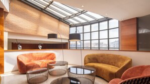 A modern interior with a high ceiling, large windows, and a brick wall. There are two sofas and three round tables. - EXE Toscana (Lucca)