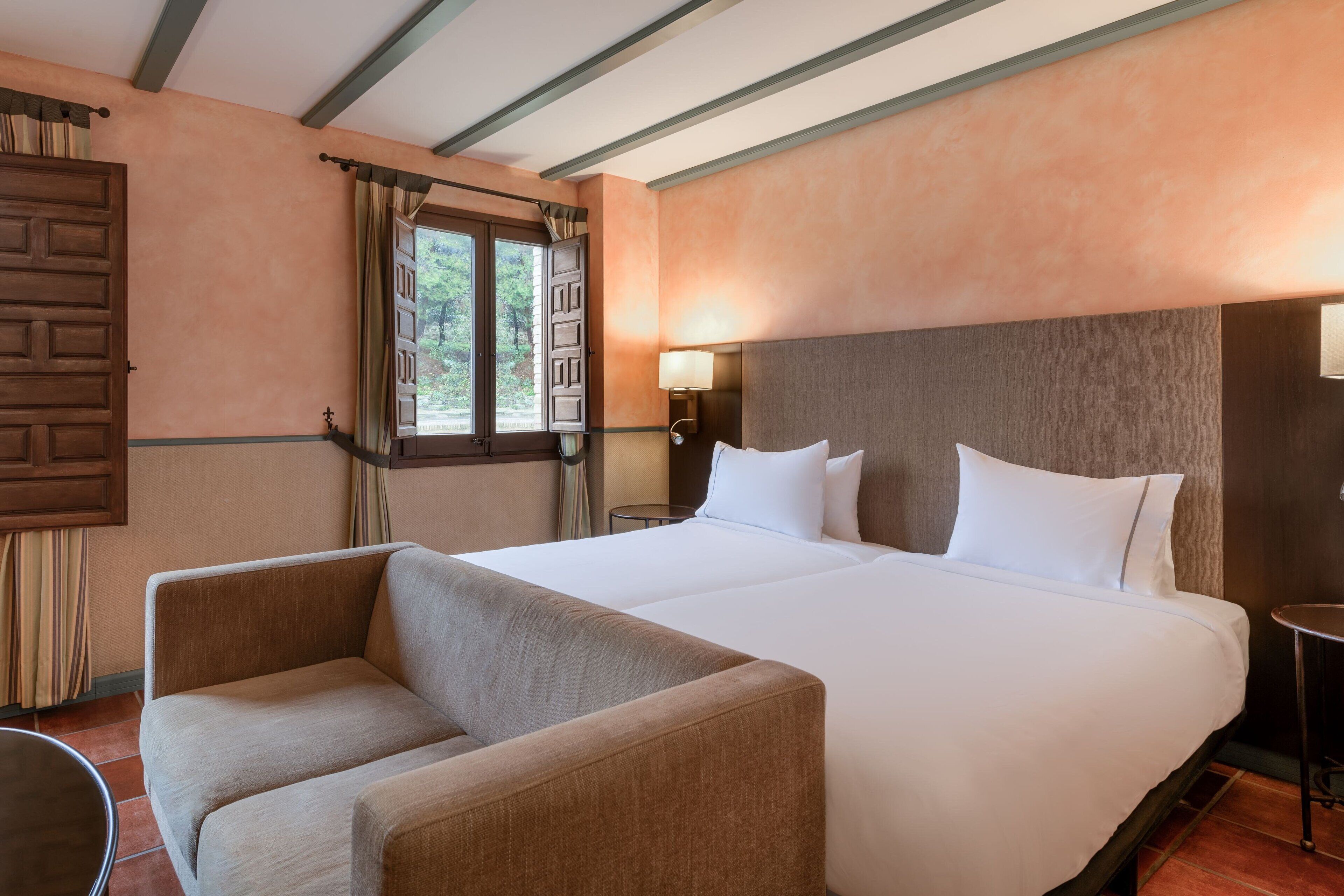 Una habitación de hotel con una cama grande, un sofá, una ventana con cortinas y una mesita de noche con lámpara.