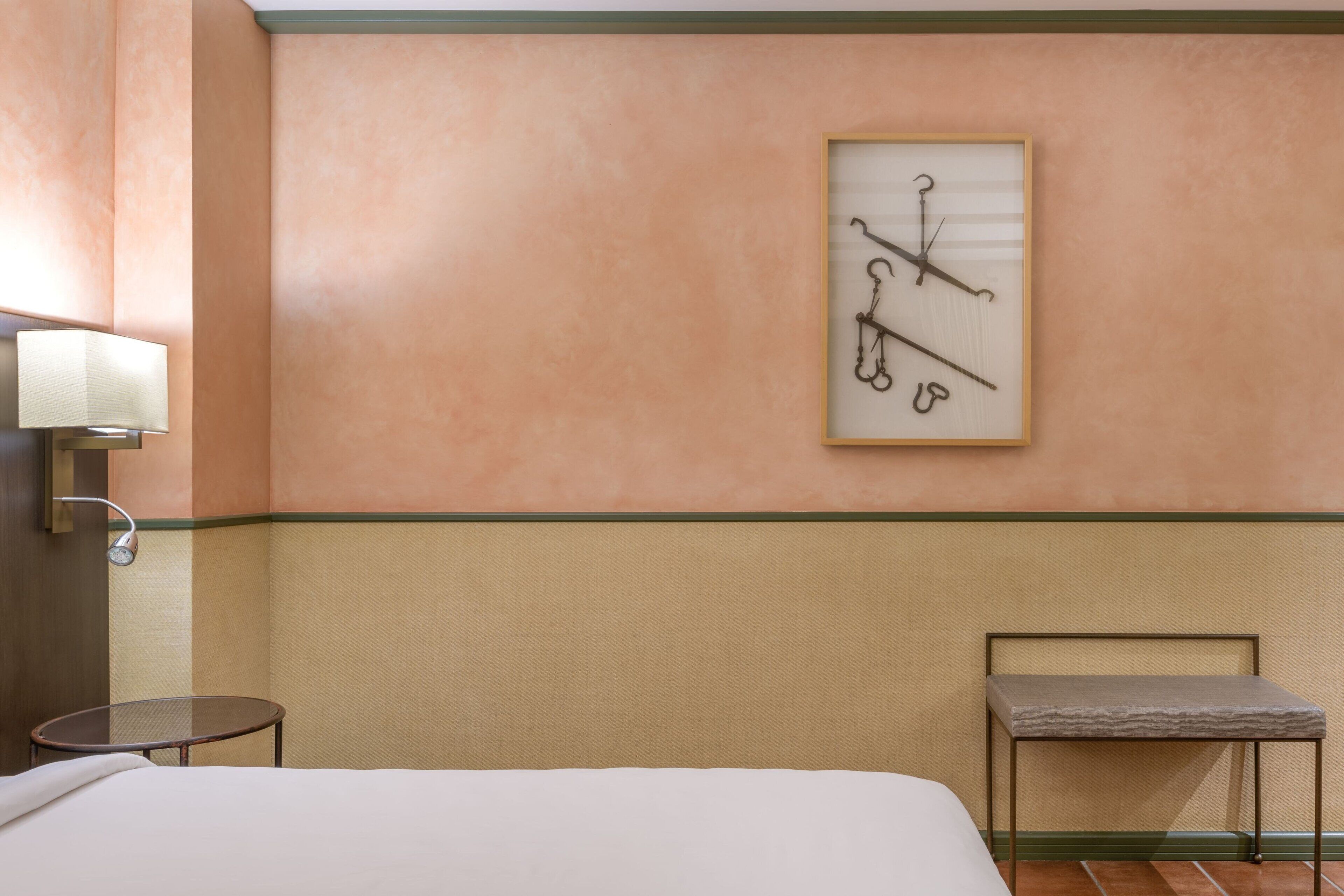 Quarto de hotel com cama, uma pequena mesa redonda, um banco, um relógio fixado na parede e uma luminária de parede.