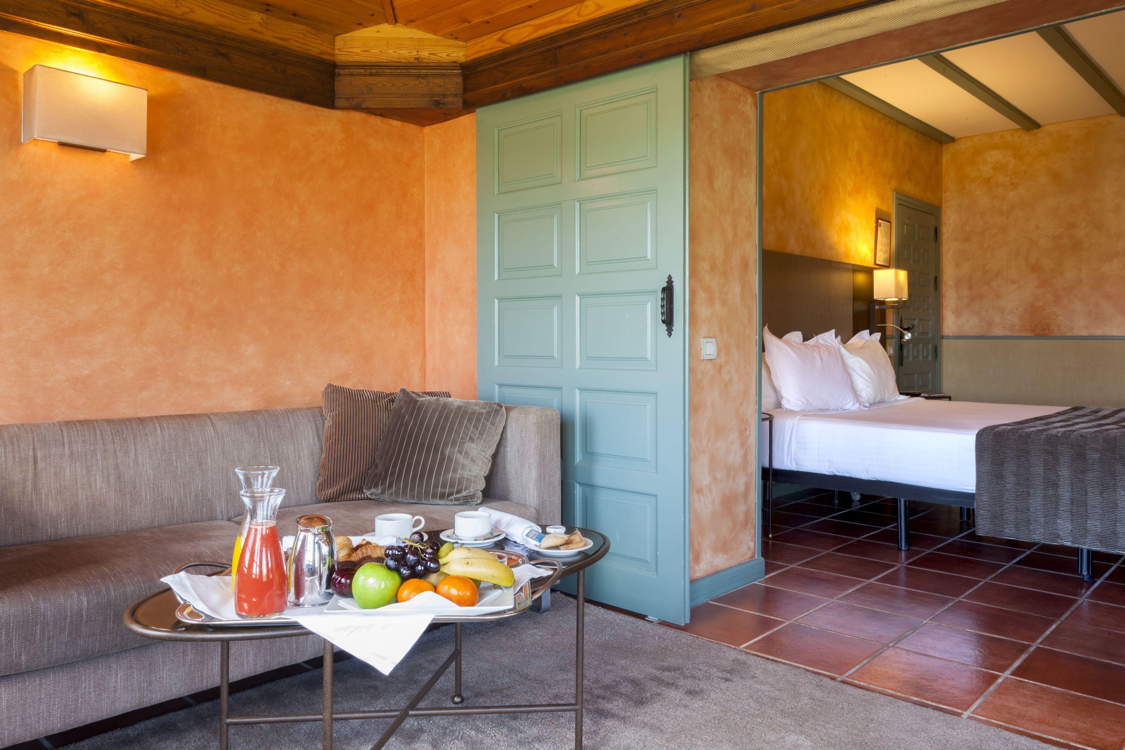 Una habitación con cama, sofá, mesa de desayuno, frutas y bebidas, y una puerta.