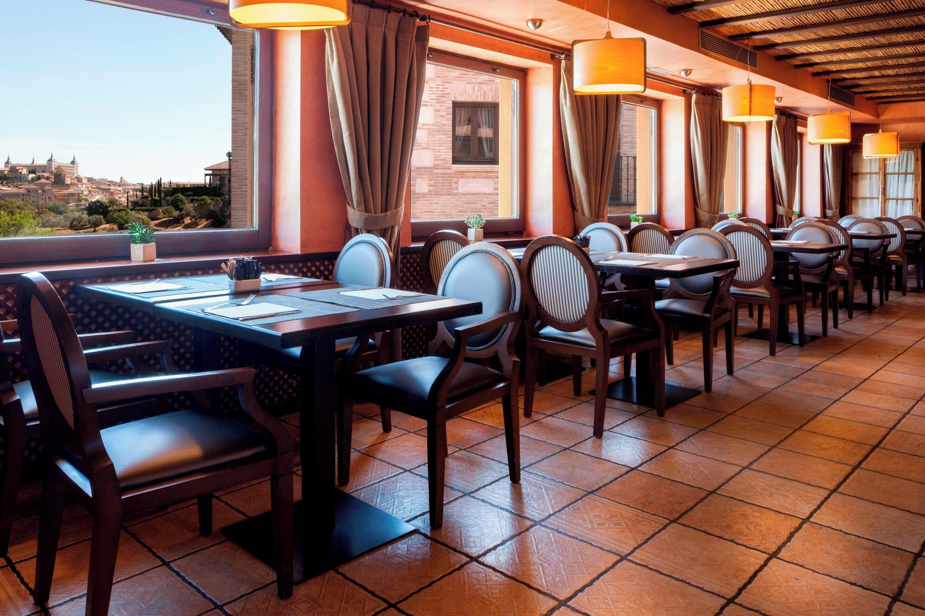 Un restaurante con varias mesas y sillas, ventanas amplias con cortinas y suelo de baldosas.