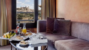 Une chambre d’hôtel dotée d’une grande fenêtre donnant sur le paysage urbain, d’un canapé, d’un plateau avec des articles pour le petit-déjeuner et d’une table sur laquelle se trouve une carafe de jus d’orange.