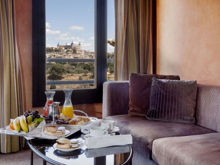 Habitación de hotel con un amplio ventanal que ofrece vistas a la ciudad, un sofá, una bandeja con alimentos para el desayuno y una mesa con jarra de jugo de naranja.