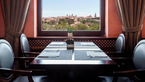 Un coin repas avec une table dressée pour deux personnes, une vue sur le paysage urbain par la fenêtre et une plante en pot sur la table.