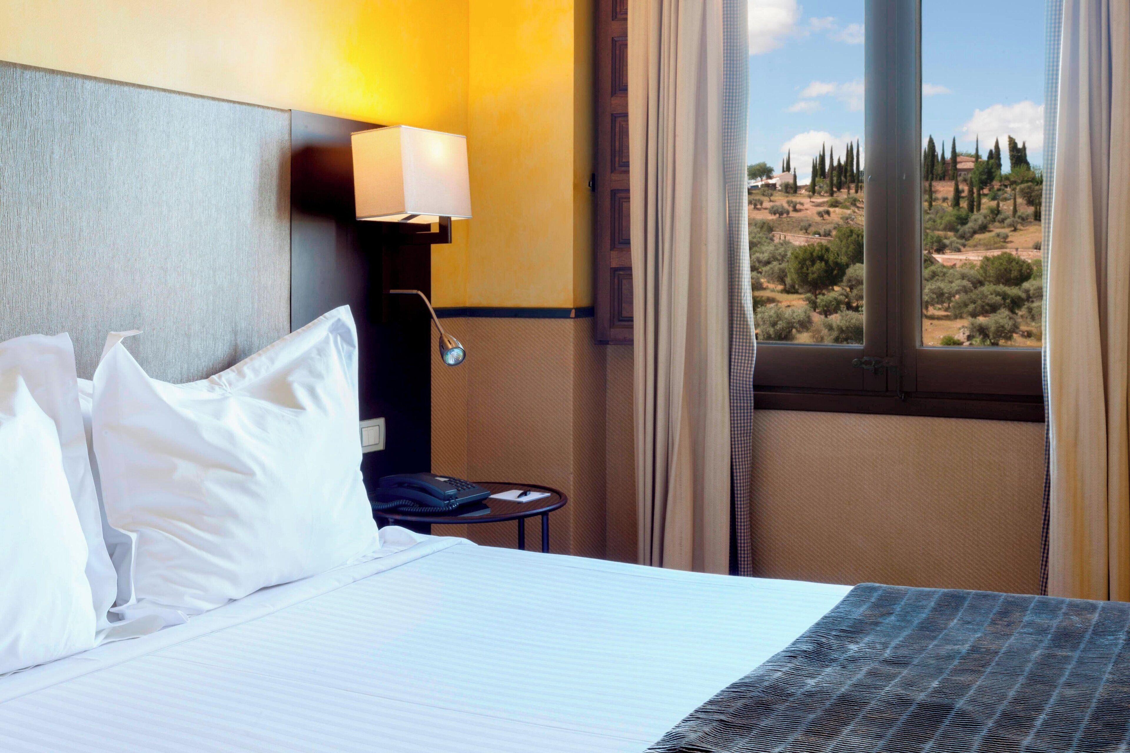 A hotel room with a bed, a bedside lamp, a telephone, and a view of a desert landscape.