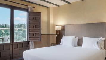 Quarto de hotel com uma cama grande, uma mesa de cabeceira com abajur, uma janela com vista para vegetação e uma porta para varanda.