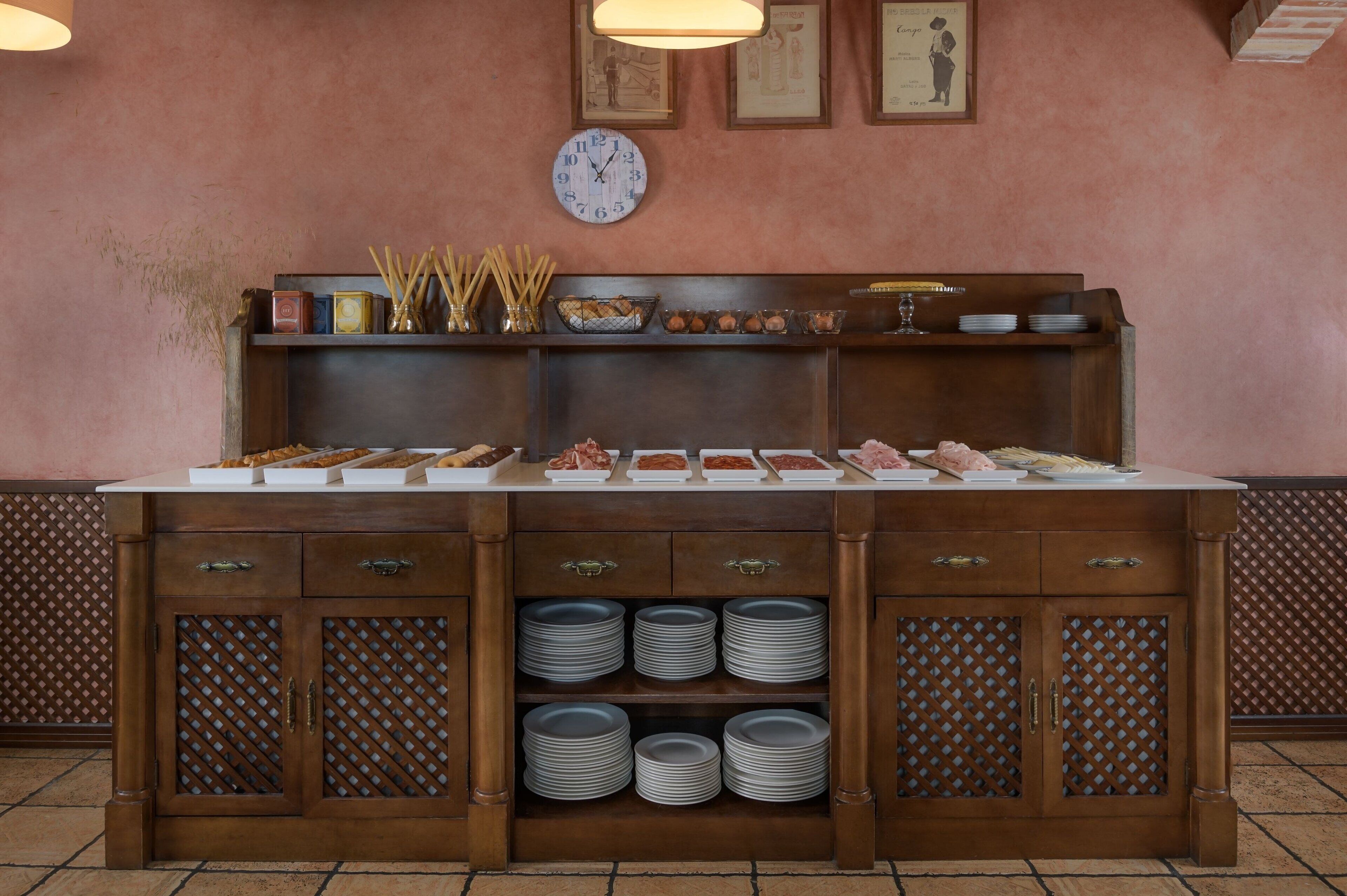 A buffet setup with various food items, a clock, and stacked plates on a wooden cabinet.