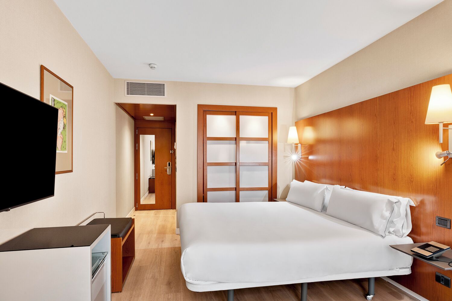 Habitación de hotel con cama, mesita de noche, televisor, armario y lámpara de pared.
