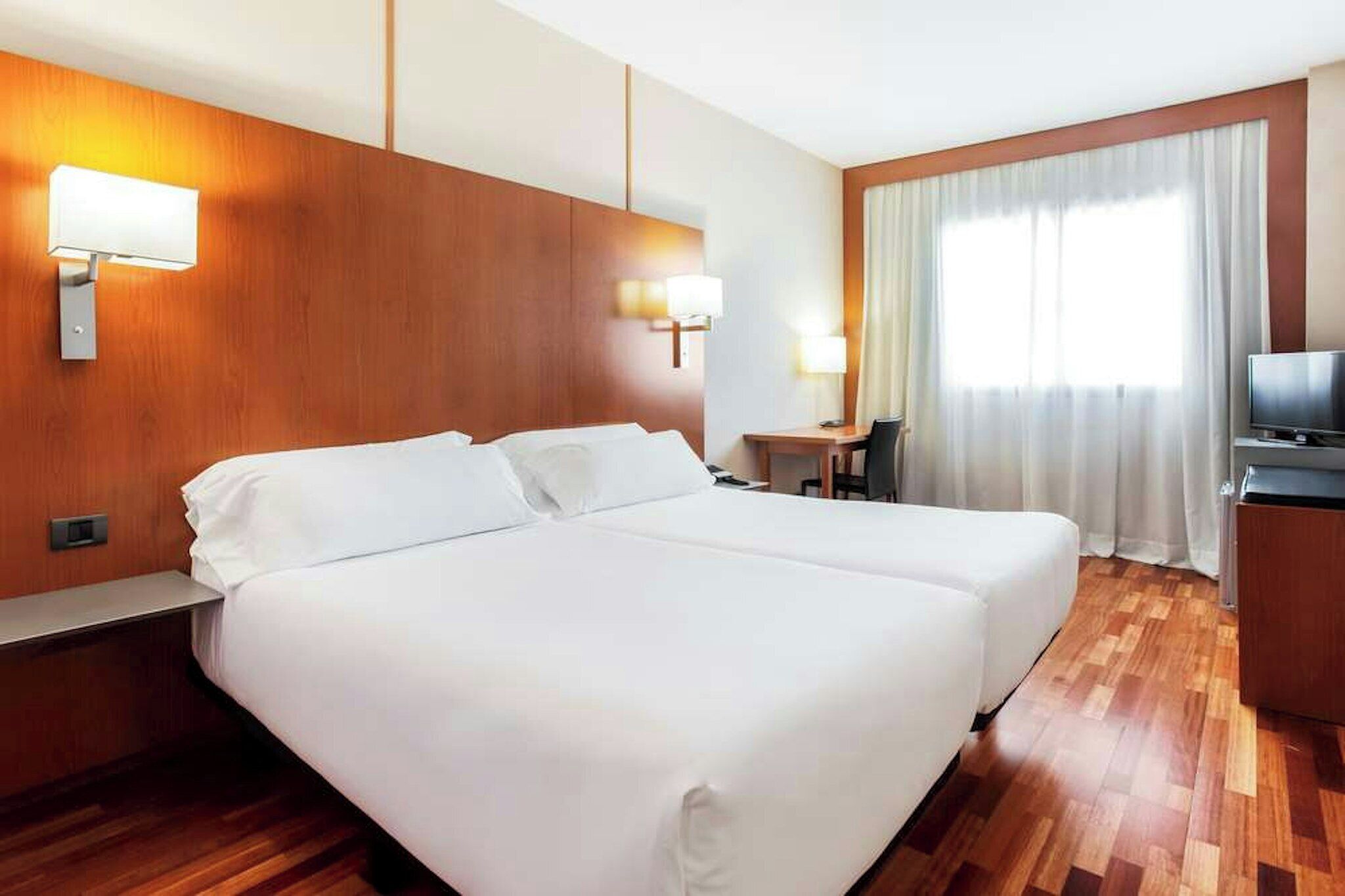 A hotel room with a double bed, a desk with a chair, a television, and a window with curtains.