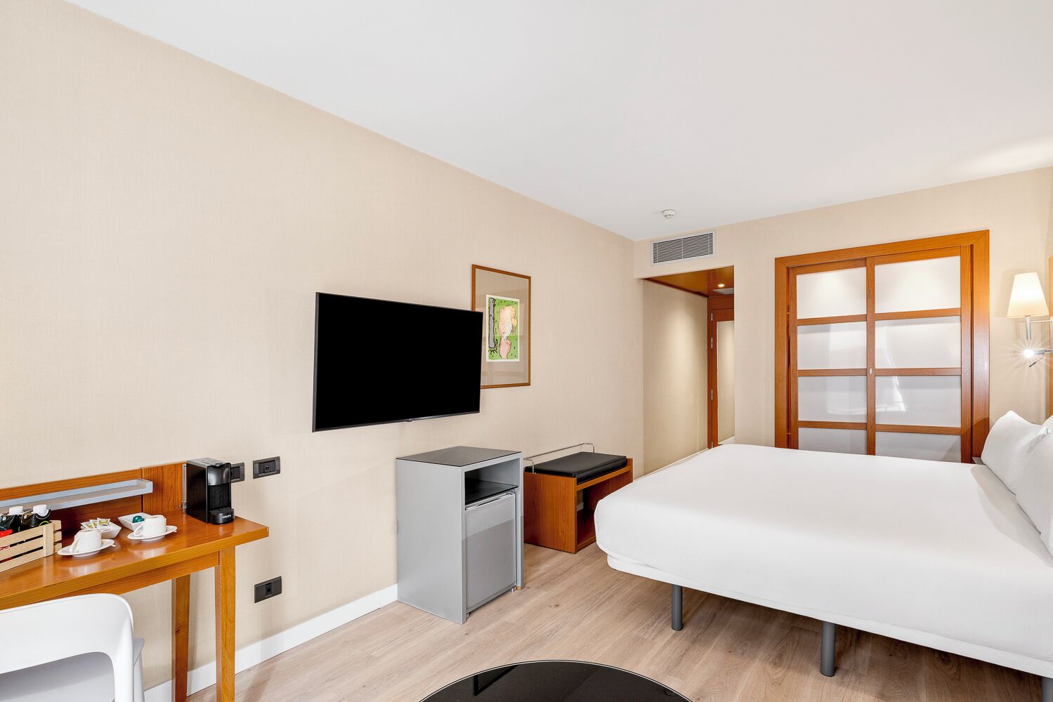 A hotel room with a bed, a desk with a coffee maker, a TV, a small fridge, and a wardrobe with a mirror.