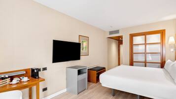 A hotel room with a bed, a desk with a coffee maker, a TV, a small fridge, and a wardrobe with a mirror.