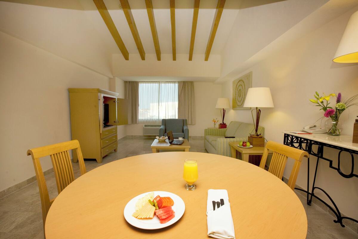 Ein Hotelzimmer mit einem runden Esstisch, einer Fruchtschale, einem Glas Orangensaft und einem ordentlich gefalteten Servietten.