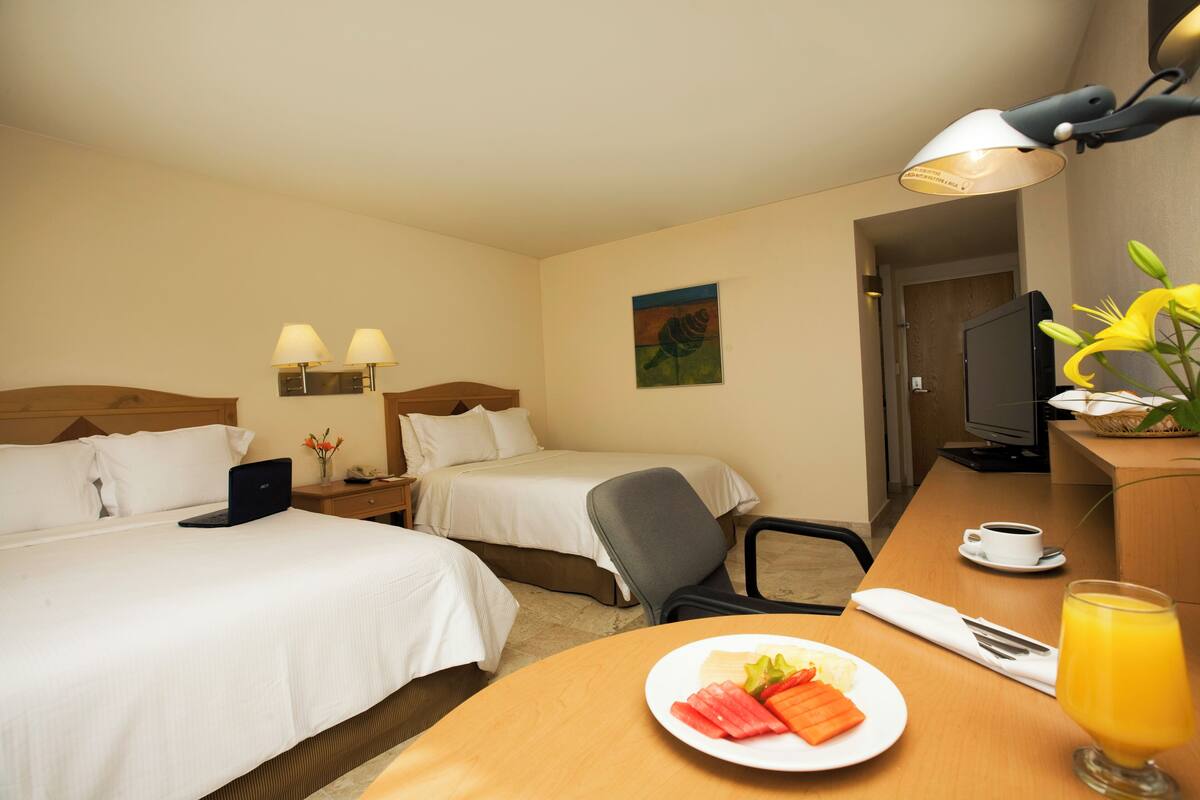 Ein Hotelzimmer mit zwei Betten, einem Schreibtisch mit einem Laptop, einem Sessel, einem Fernseher und einem Frühstückswagen mit Obst und Saft.