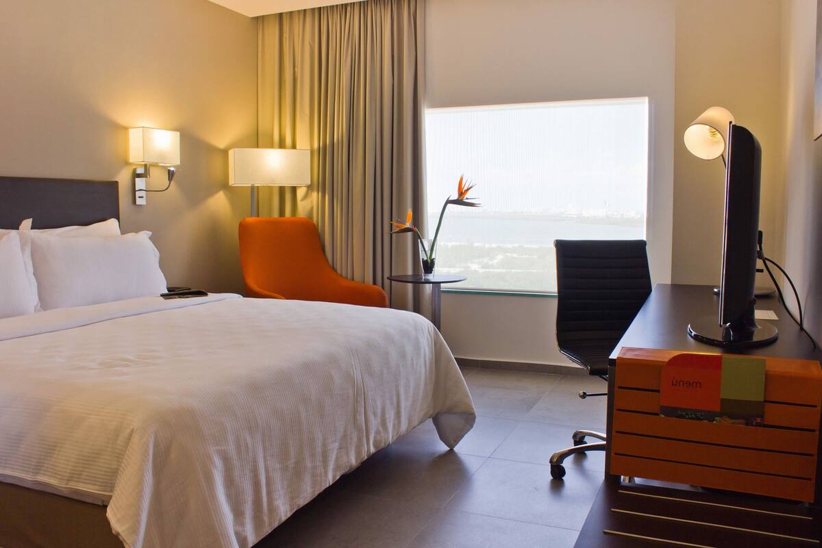 Ein Hotelzimmer mit einem großen Bett, einem orangefarbenen Sessel, einem Schreibtisch mit Computer und Blick auf die Stadt durch ein Fenster.
