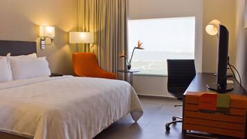 A hotel room with a large bed, an orange chair, a desk with a computer, and a view of the city through a window.