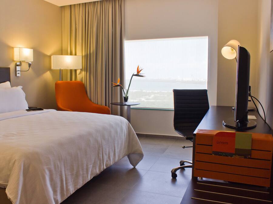 A hotel room with a large bed, an orange chair, a desk with a computer, and a view of the city through a window.