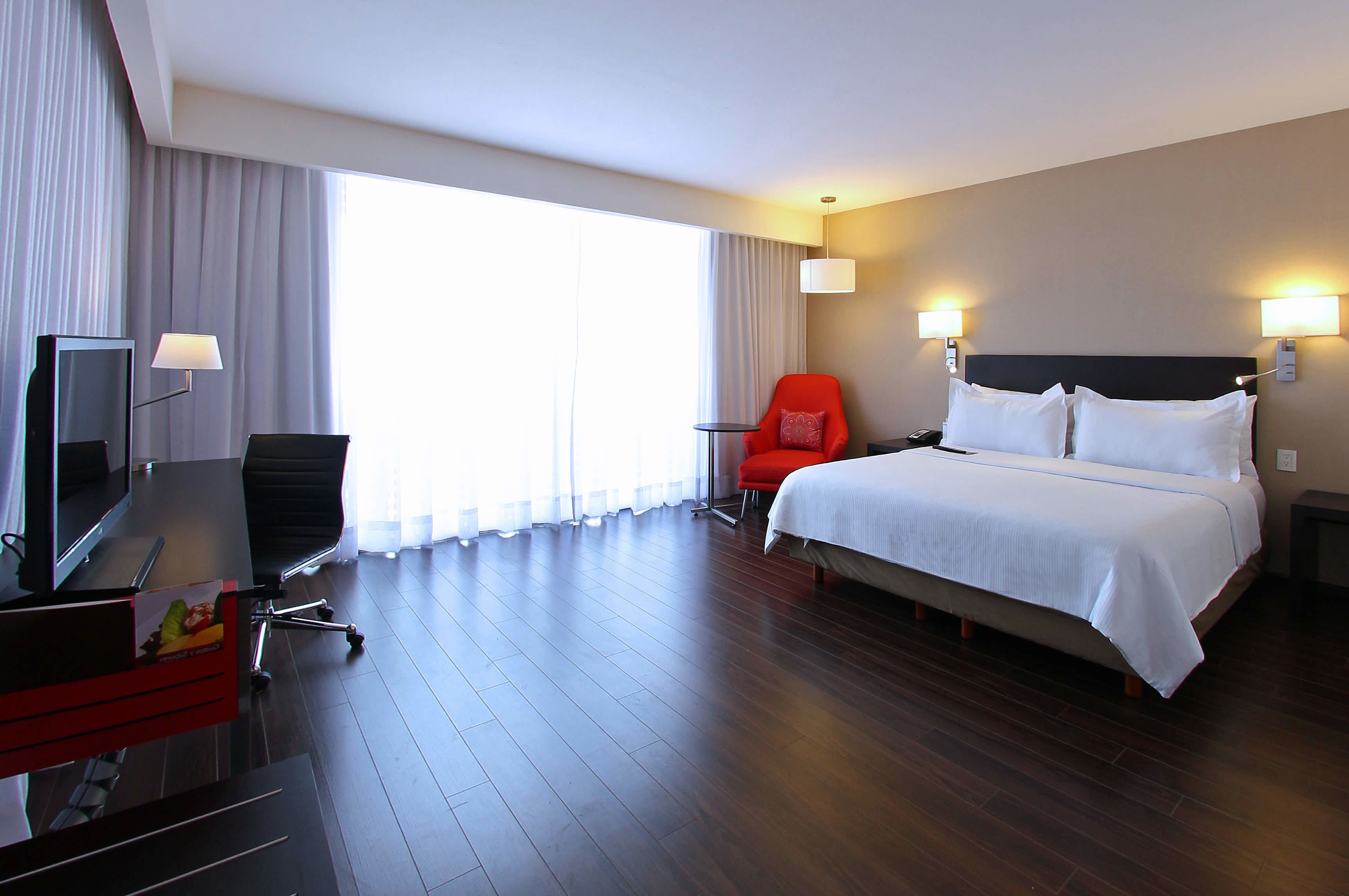Une chambre d’hôtel avec un lit, un bureau avec un ordinateur, une chaise rouge et un téléviseur.