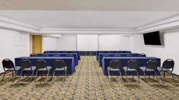 A conference room with rows of tables and chairs, a carpeted floor, and a flat-screen TV on the wall.
