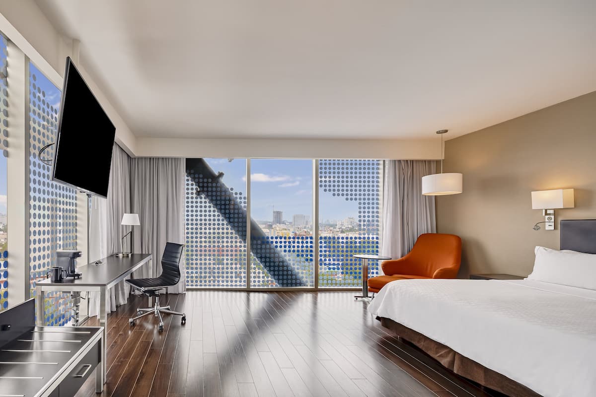 Habitación de hotel con una cama grande, un escritorio con silla, televisión y vistas a la ciudad a través de amplios ventanales.