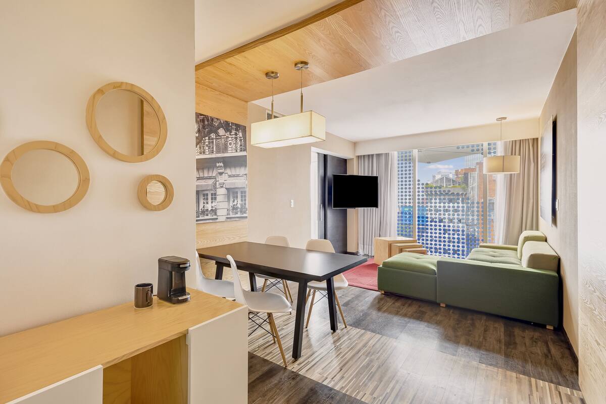 Habitación de hotel moderna con zona de comedor, sofá, televisor y vista a la ciudad.