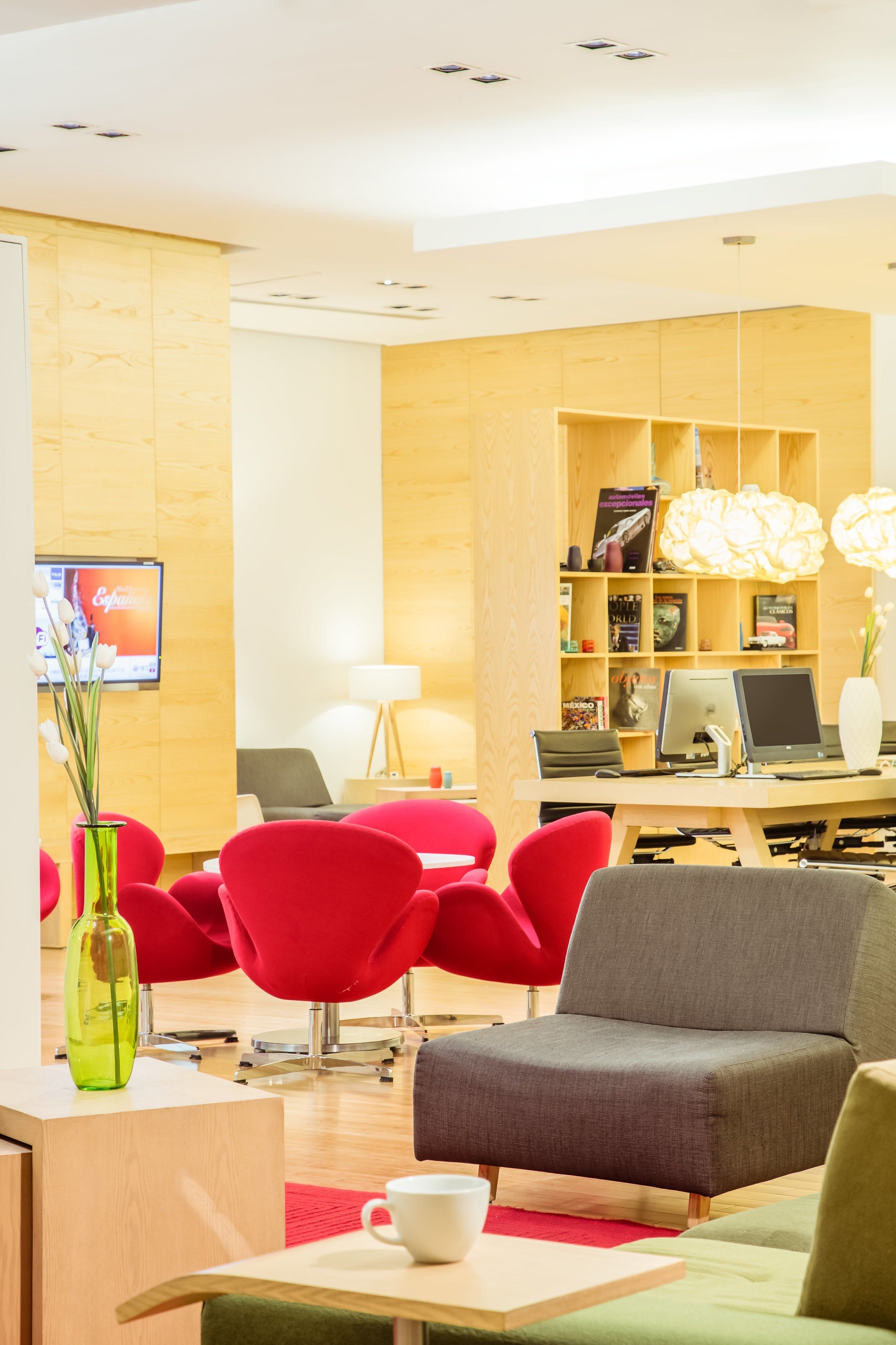 A modern reception area with a wooden desk, red chairs, a grey sofa, and a bookshelf with books and a computer.