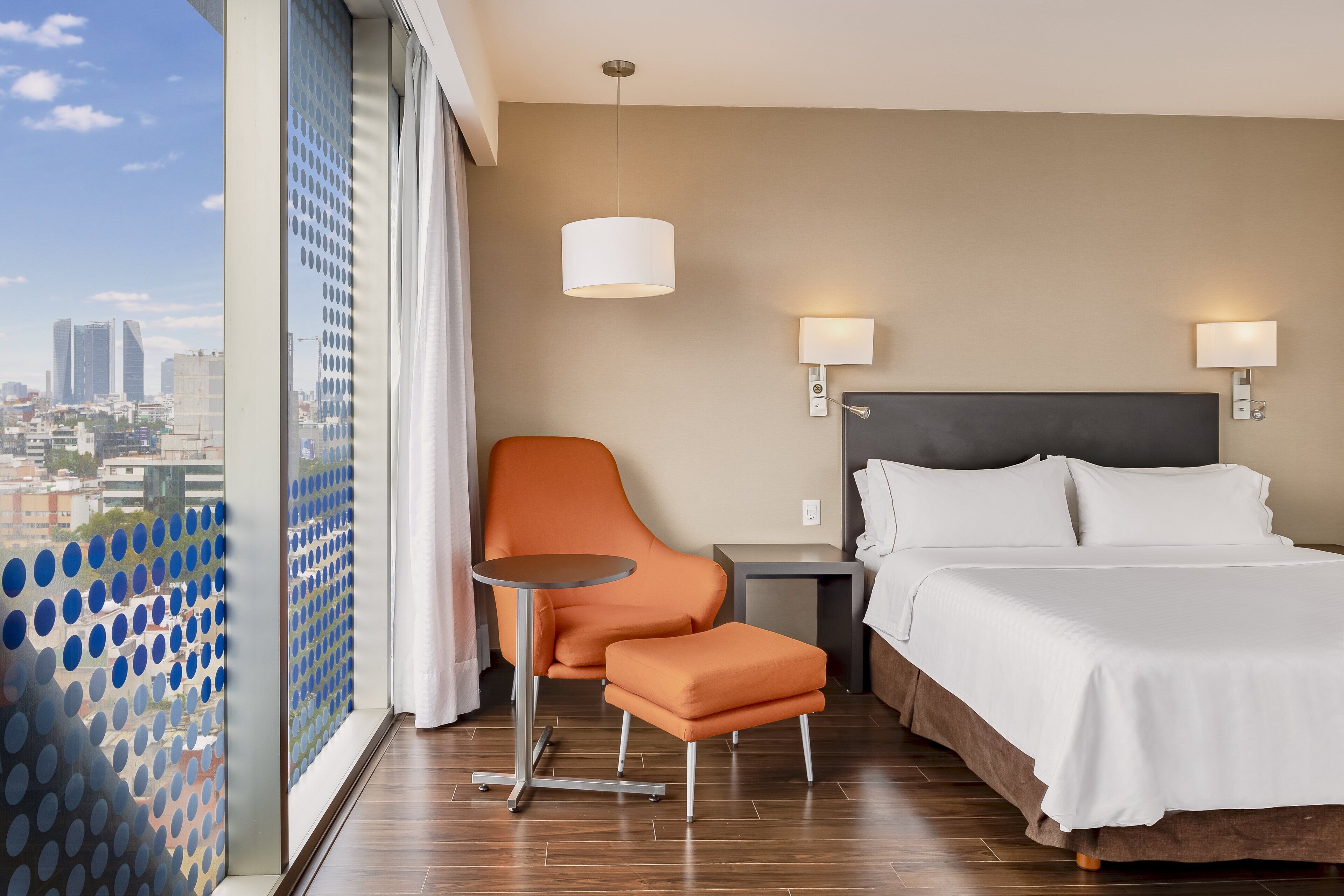 Een hotelkamer met een bed, een oranje stoel, een klein tafeltje en uitzicht op de stad door een raam met een blauw stippatroon.