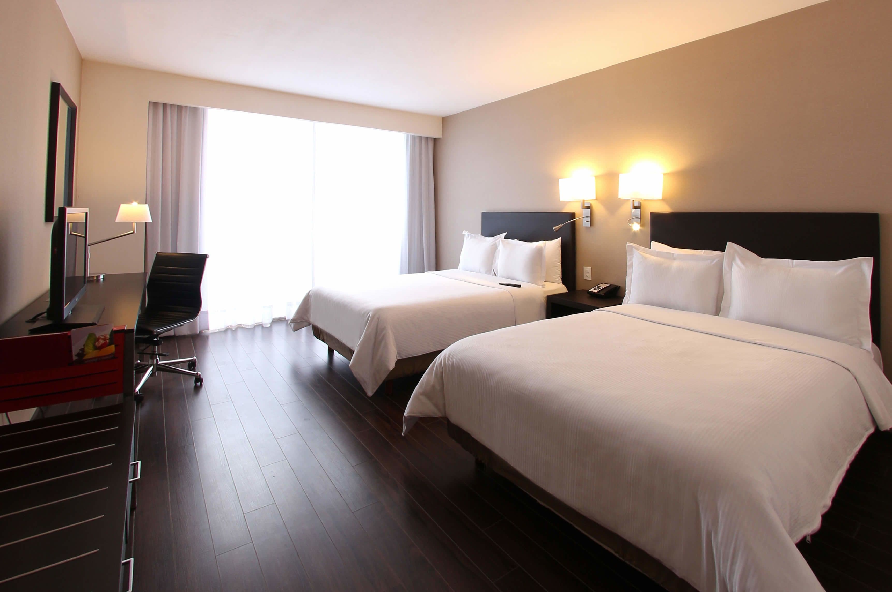 A hotel room with two beds, a desk, a chair, and a large window with curtains.