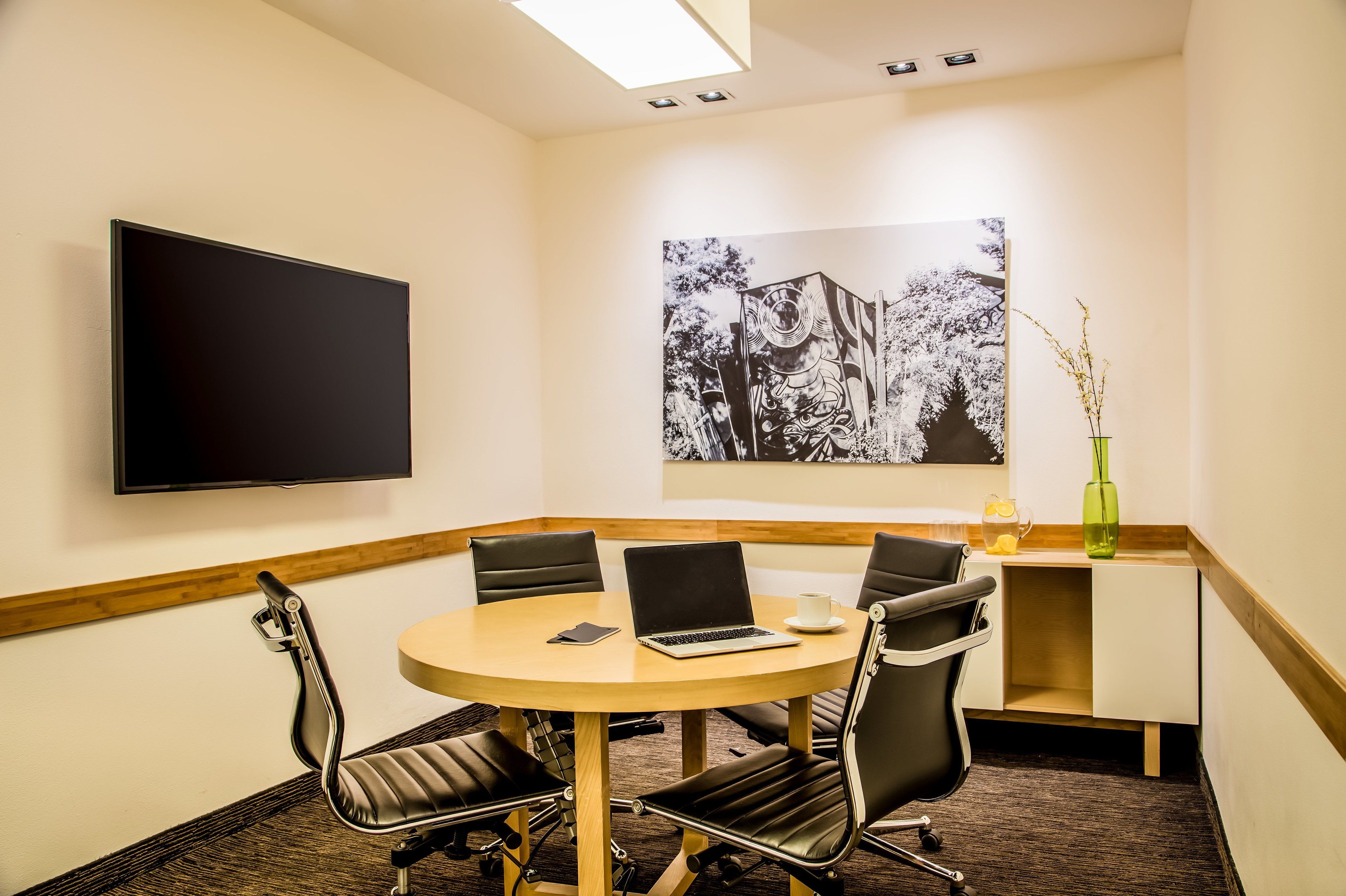 A conference room with a round table, four chairs, a laptop, a cup, and a vase with branches.