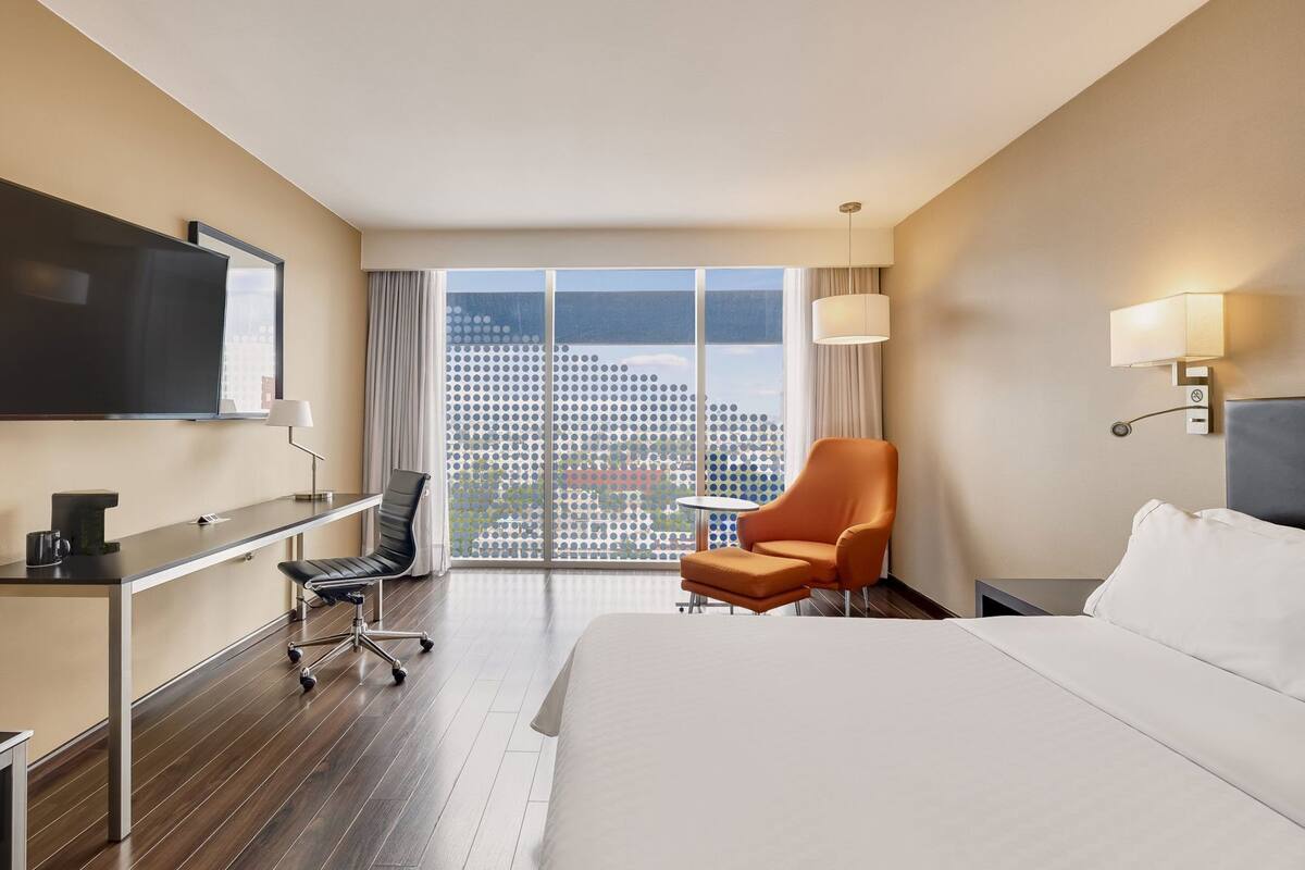 Habitación de hotel con cama, escritorio, silla, televisión y un amplio ventanal con vistas a edificios.
