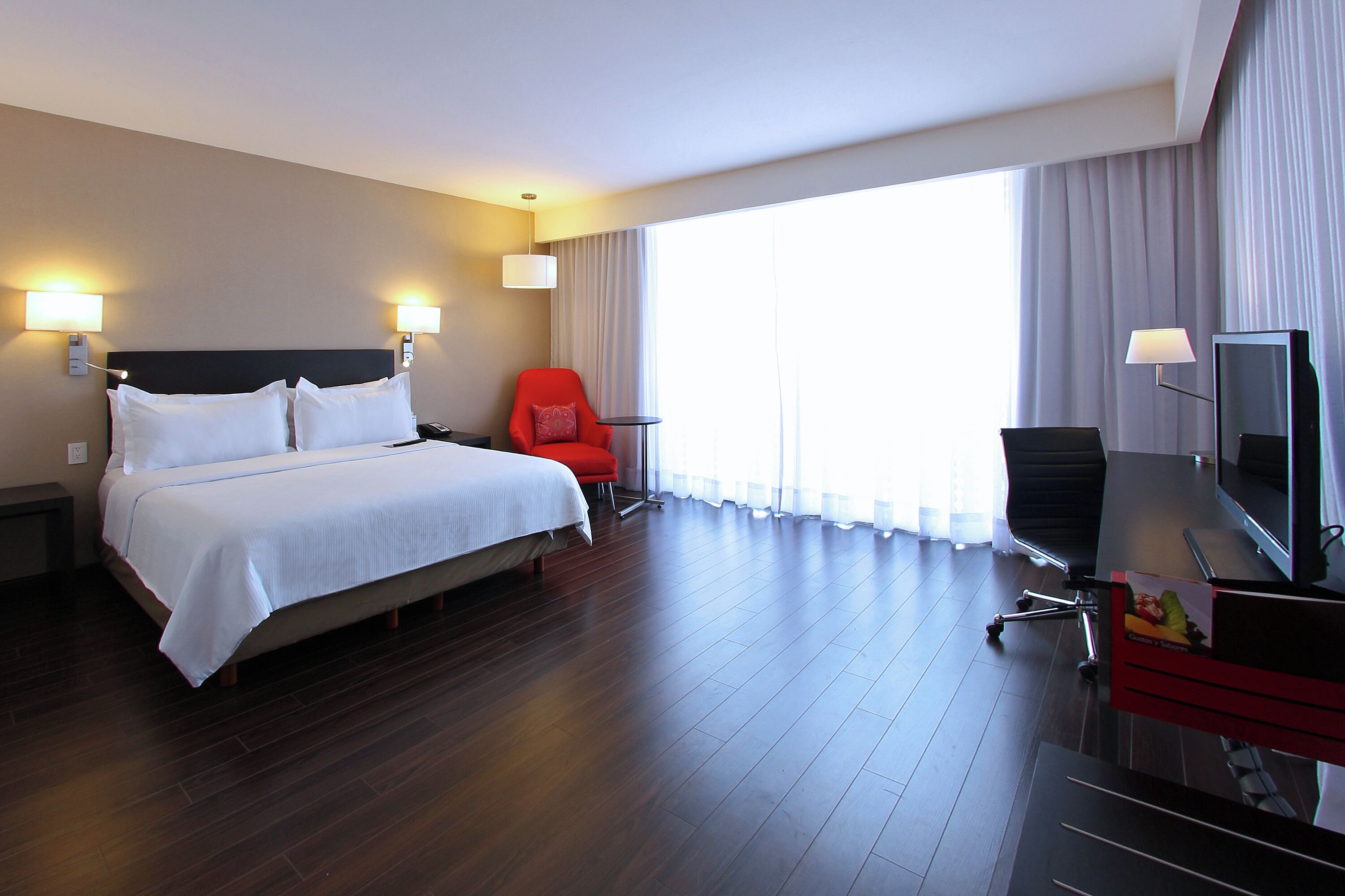 Une chambre d’hôtel avec un grand lit, deux lampes de chevet, un fauteuil rouge, un bureau avec une télévision et une grande fenêtre avec des rideaux.