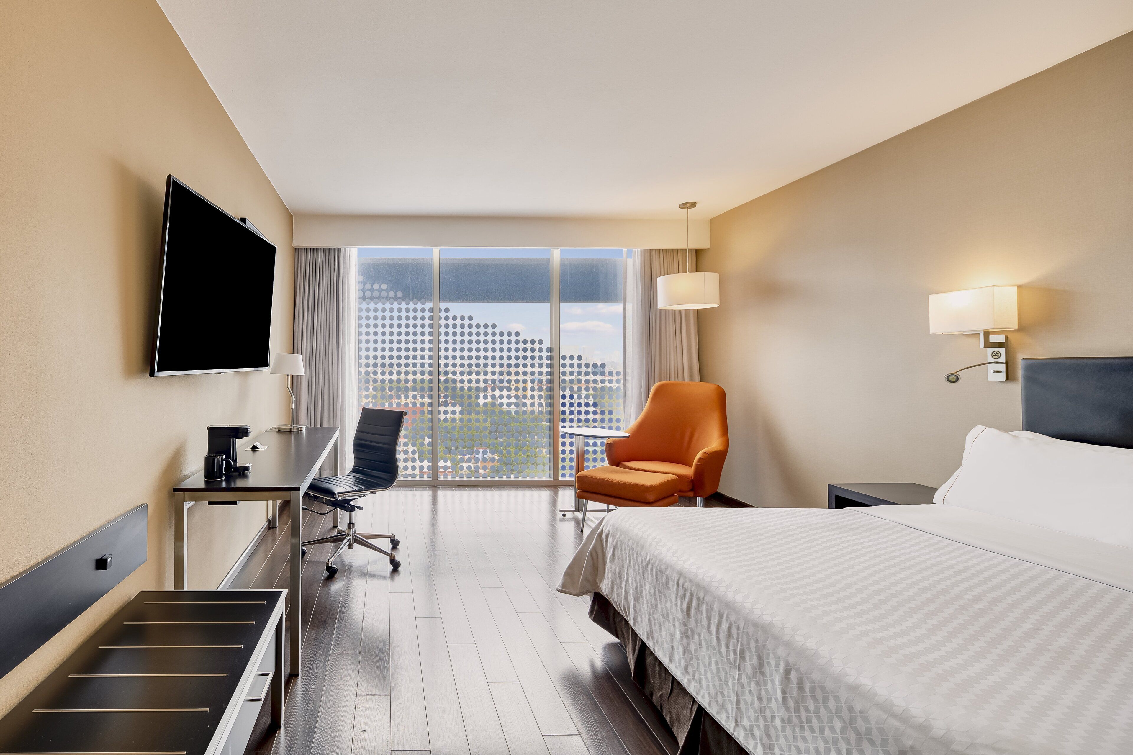 Una habitación de hotel con una cama, un escritorio con silla, un televisor y un ventanal con vistas a la ciudad.
