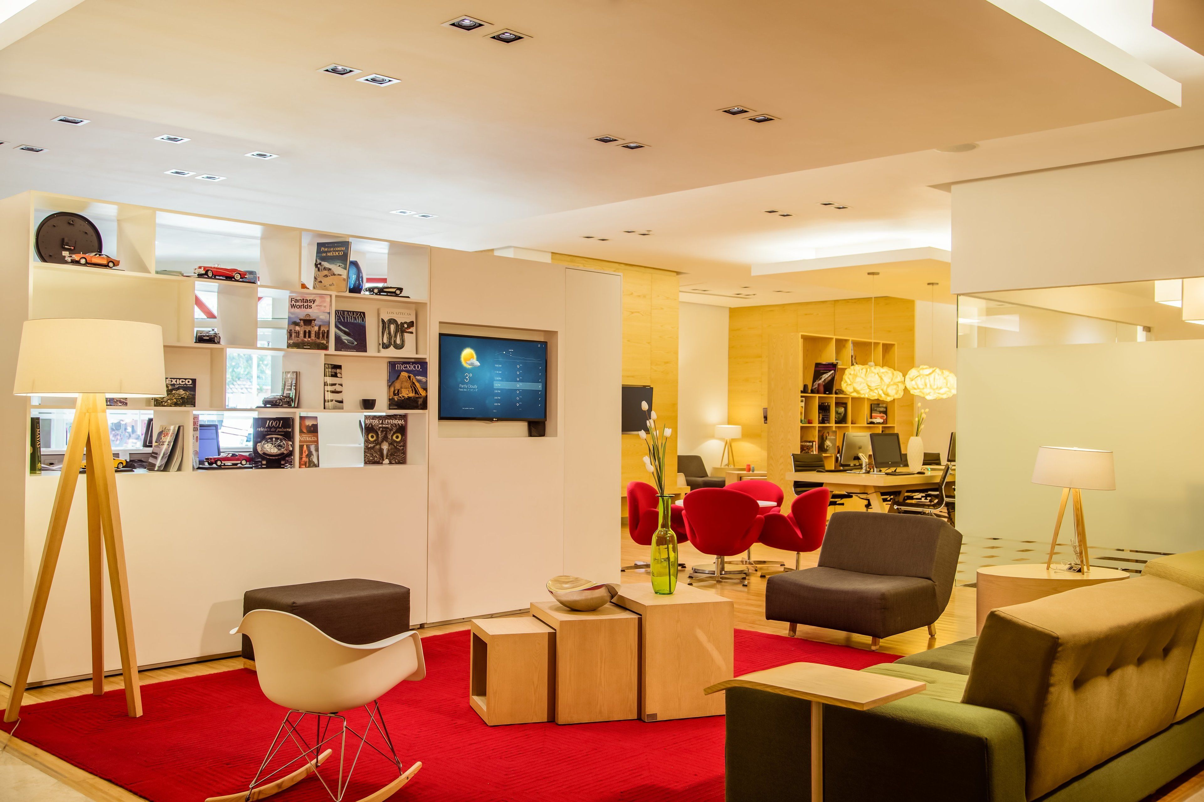 Un hall d’hôtel moderne avec un tapis rouge, des espaces aménagés pour s’asseoir et une bibliothèque contenant divers livres et objets.