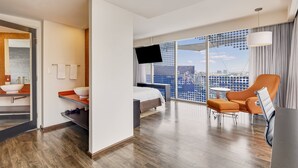 A modern hotel room with a bed, a TV, a seating area with a chair and ottoman, a small table, and a large window with a city view. - Fiesta Inn Insurgentes Viaducto (Mexico City)