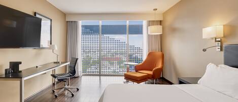 Una habitación de hotel con una cama grande, un escritorio con silla, un televisor, una ventana con vistas a un paisaje urbano y una lámpara.