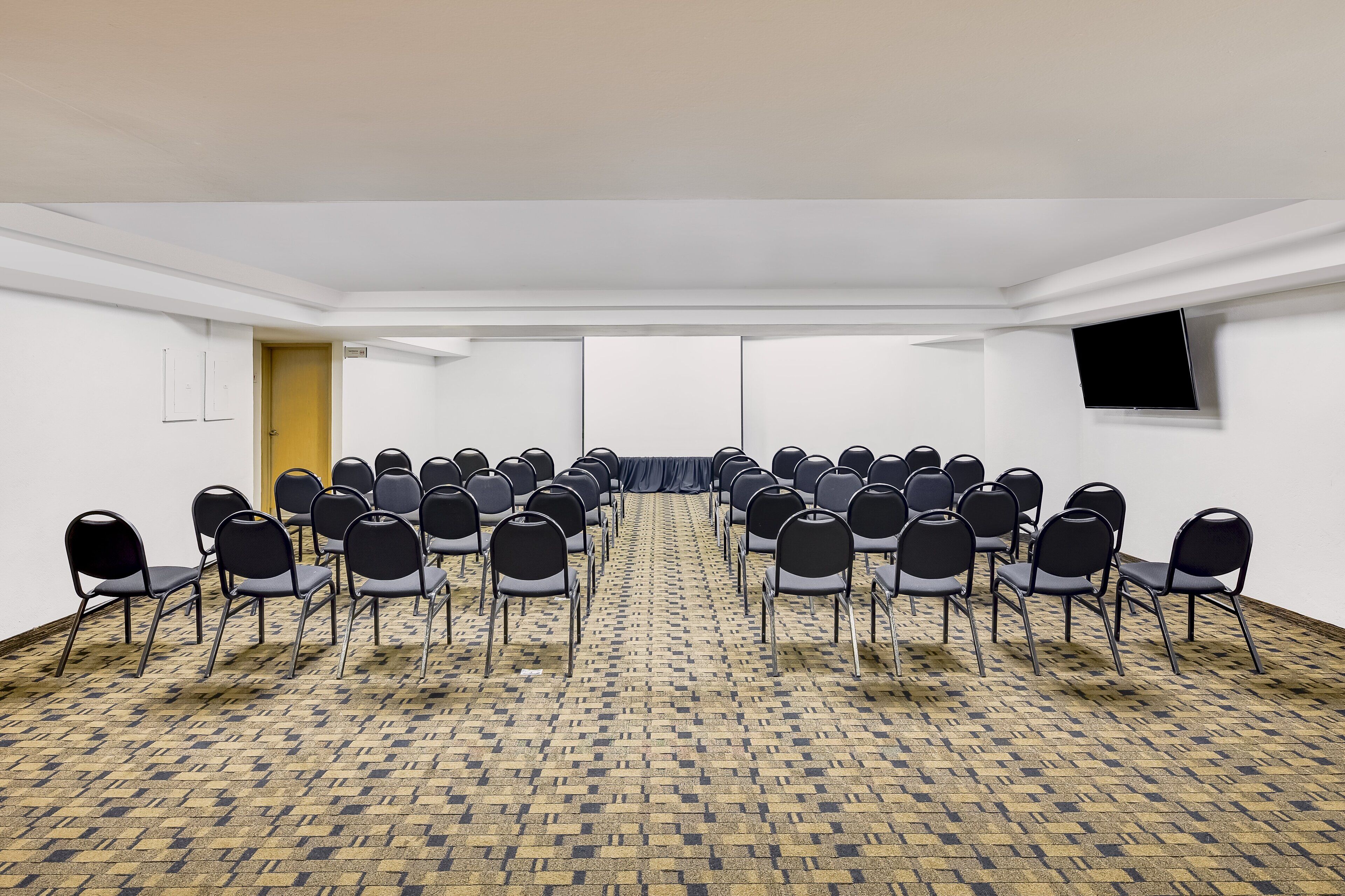 A conference room with rows of chairs, a carpeted floor, a door, and a flat-screen TV on the wall.