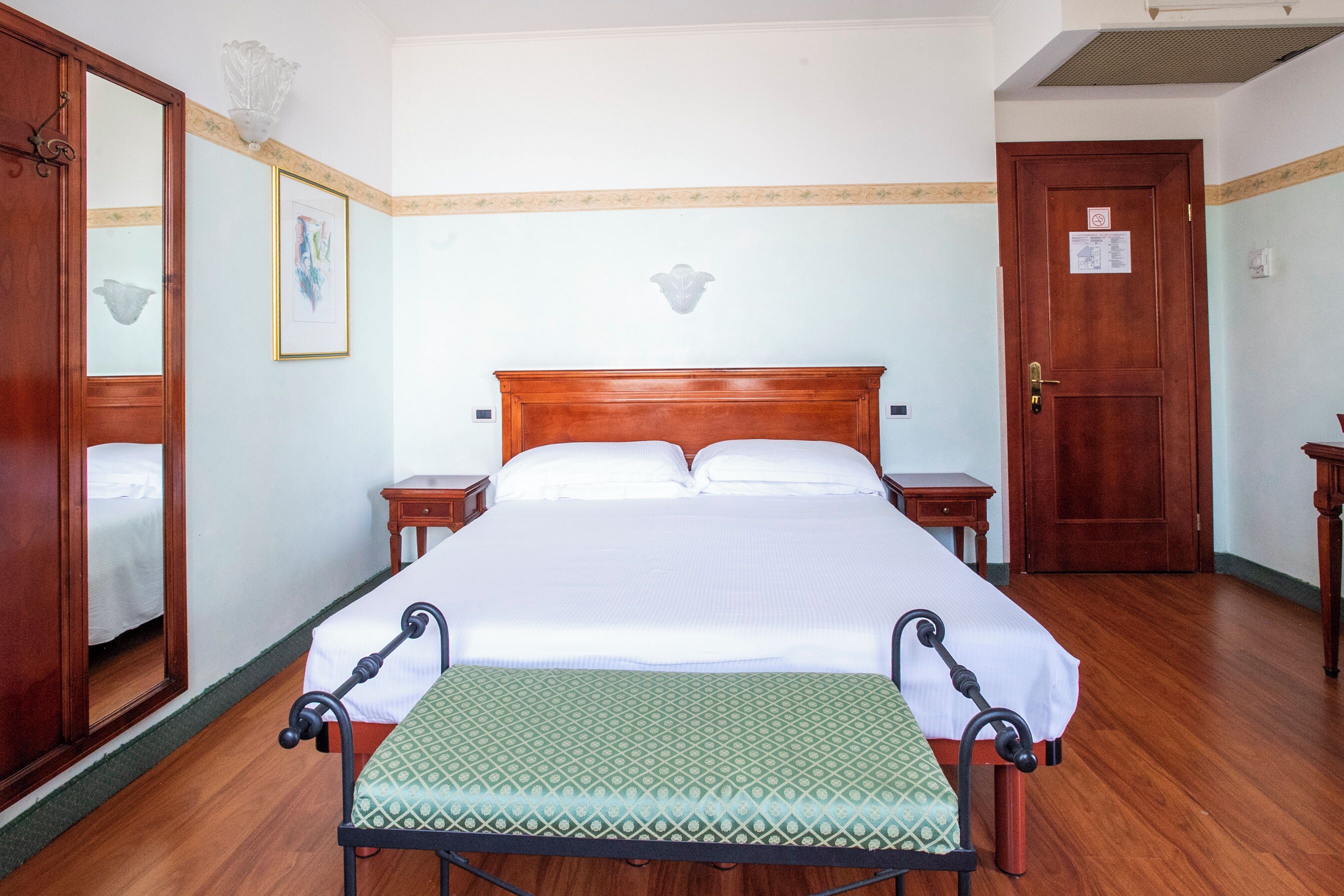 Un dormitorio ordenado con una cama de madera, mesitas de noche, un espejo y un banco verde con estampado.