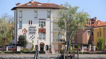 Russo Palace Hotel features a classic entrance with a decorative archway and balcony, situated beside a serene body of water.
