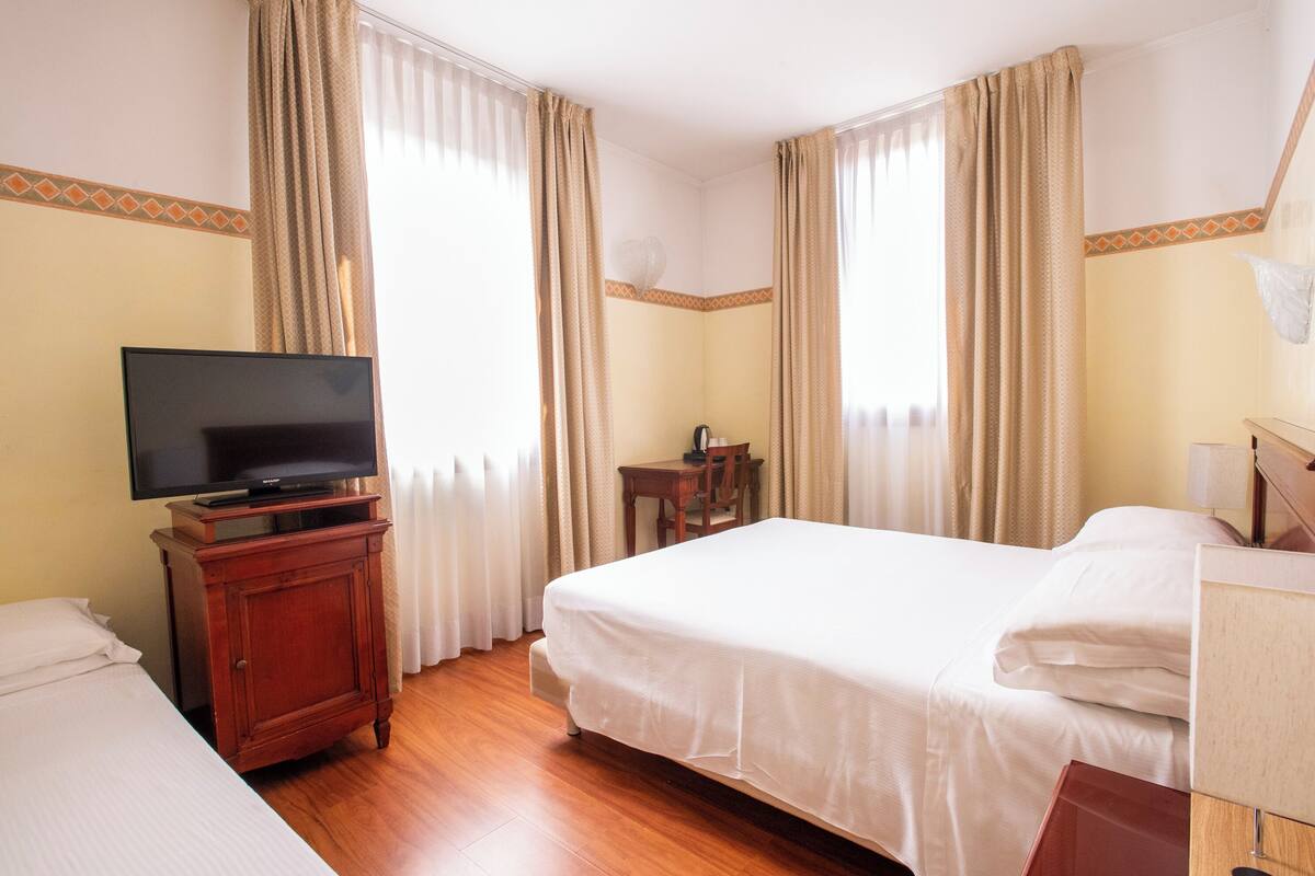 A hotel room with a bed, a TV on a wooden cabinet, a desk with a chair, and a window with curtains.