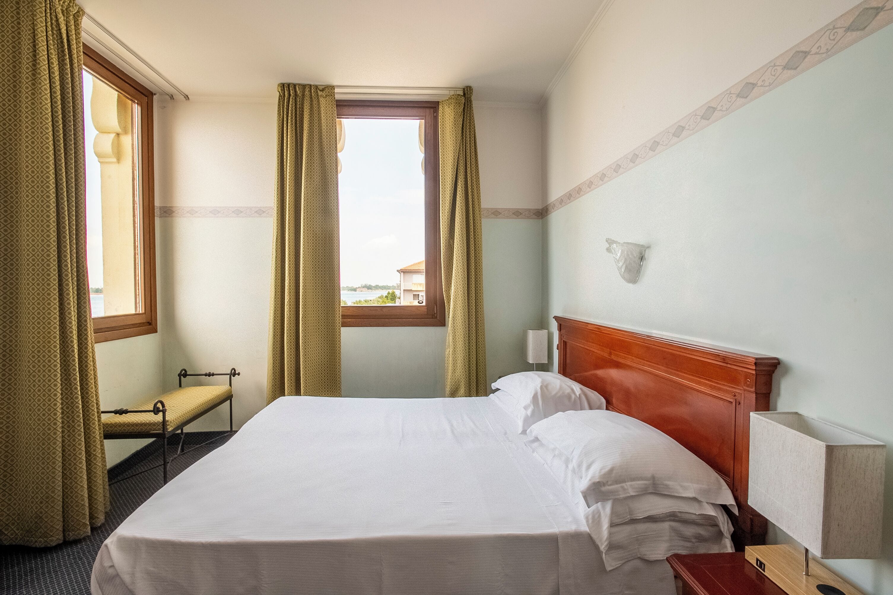 Habitación de hotel con una cama grande, un banco, una mesita de noche, una lámpara y una ventana con cortinas.