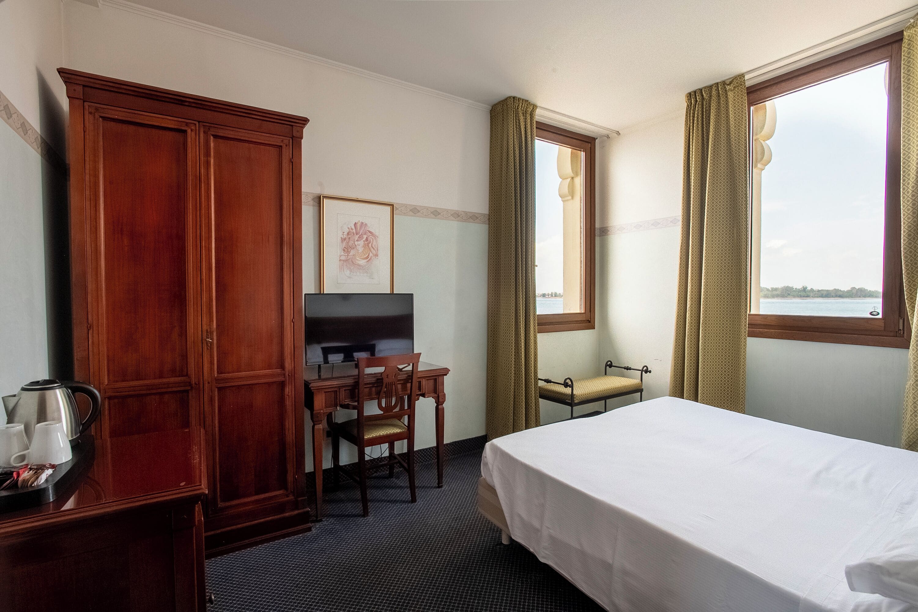 Habitación de hotel con cama, escritorio con silla, armario, ventana con cortinas e imagen enmarcada en la pared.