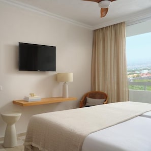 Habitación de hotel con una cama, televisión montada en la pared, mesita de noche con lámpara y vistas a la ciudad por la ventana.