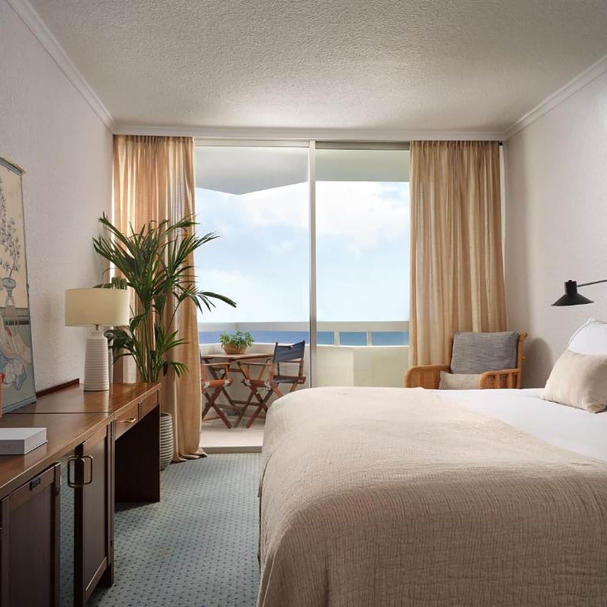 A hotel room with a large bed, a desk with a lamp, a chair, a small table with a plant, and a view of the sea through the balcony doors.