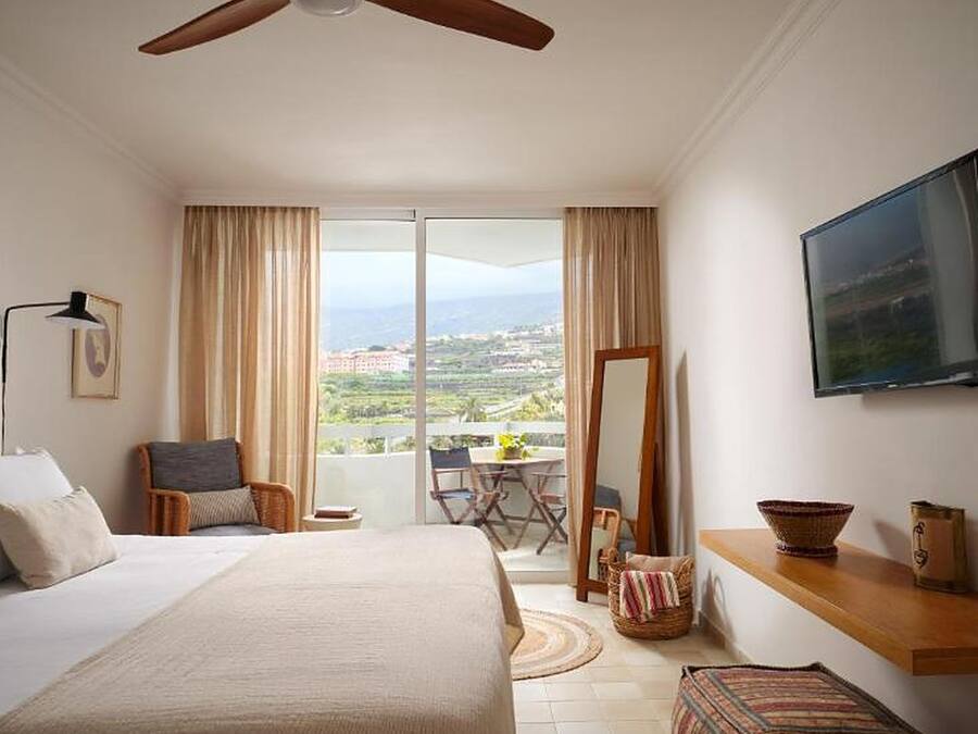 Habitación de hotel con cama, ventilador de techo, televisión, balcón con mesa y sillas, y vista del paisaje.