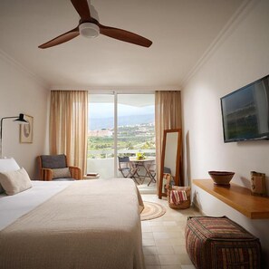 Een hotelkamer met een bed, een plafondventilator, een televisie, een balkon met een tafel en stoelen, en uitzicht op het landschap.