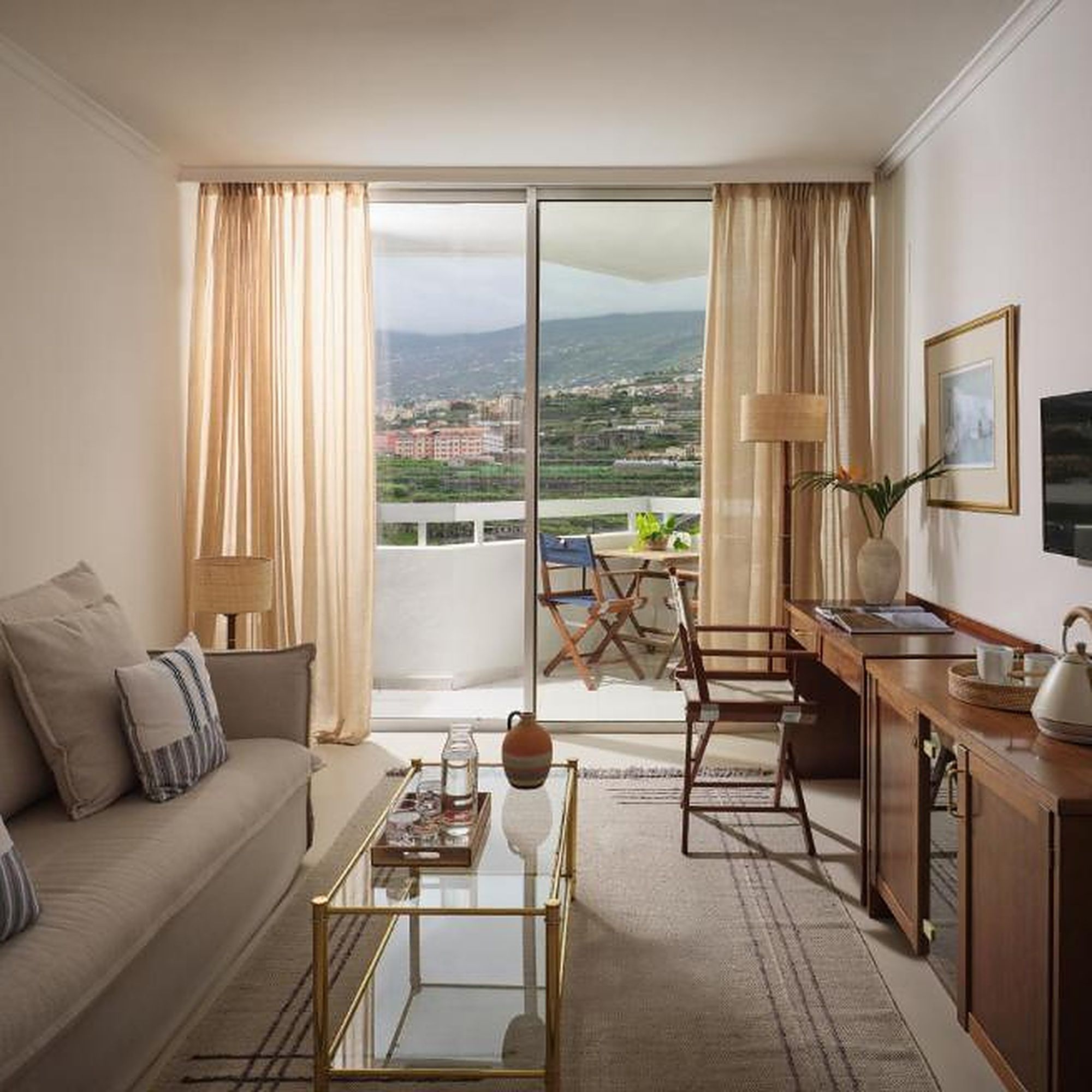 Habitación de hotel con sofá, una mesa de centro, escritorio con lámpara, vista de la ciudad a través de un ventanal grande y balcón con mobiliario exterior.