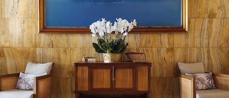 Een kamer met een groot schilderij van een berglandschap, twee gestoffeerde fauteuils, een houten console met een vaas witte bloemen en een kamerbreed tapijt.
